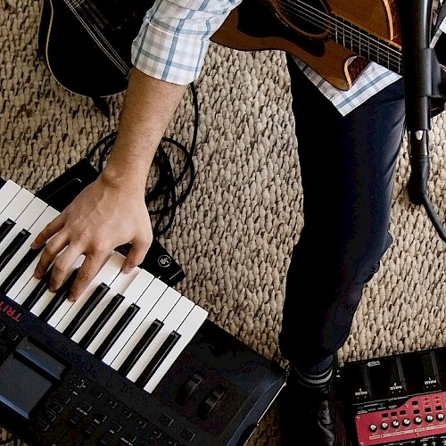 A musician is playing electronic and acoustic instruments, including a keyboard, guitar, and drum machine, on a carpeted floor.