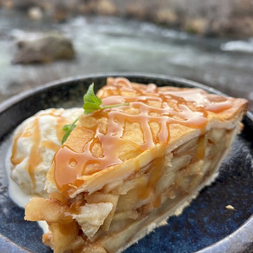 A piece of apple pie with caramel drizzle and whipped cream is served outdoors near a river.