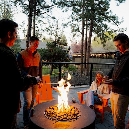 Four friends enjoy a campfire outdoors at dusk, sitting and standing around, with smiles and laughter.