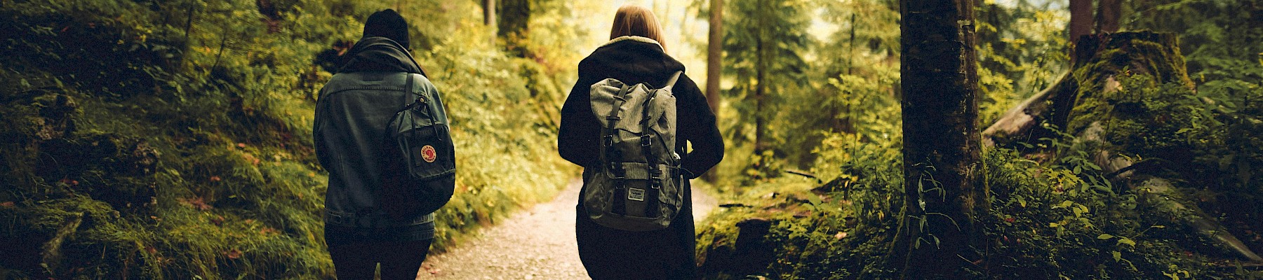 Two people with backpacks walk along a shaded forest trail during daytime.