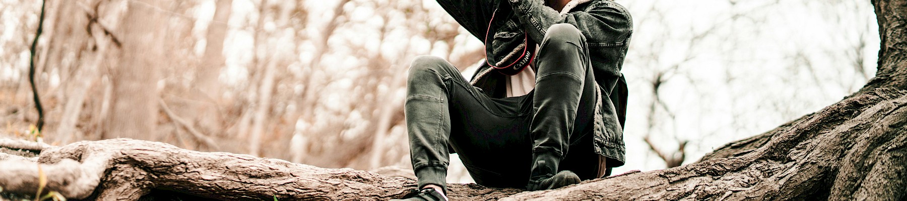 A person is sitting on tree roots in a forest, taking a photo with a DSLR camera.