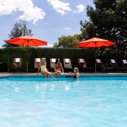 Three women sit by a swimming pool with red umbrellas, chatting and relaxing on poolside chairs on a sunny day.