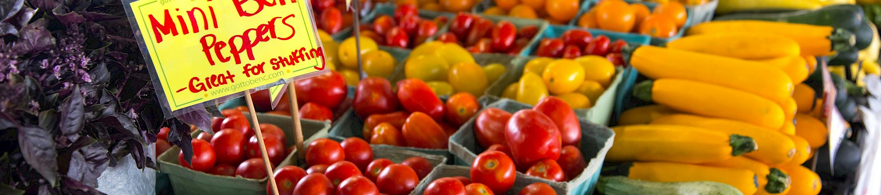 A market stand displays colorful fresh vegetables and a sign promoting mini bell peppers.