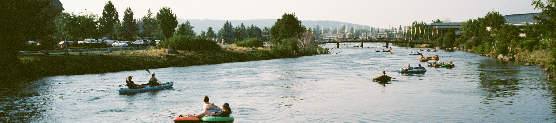 People are kayaking and paddleboarding on a river under a clear sky with greenery on the banks.
