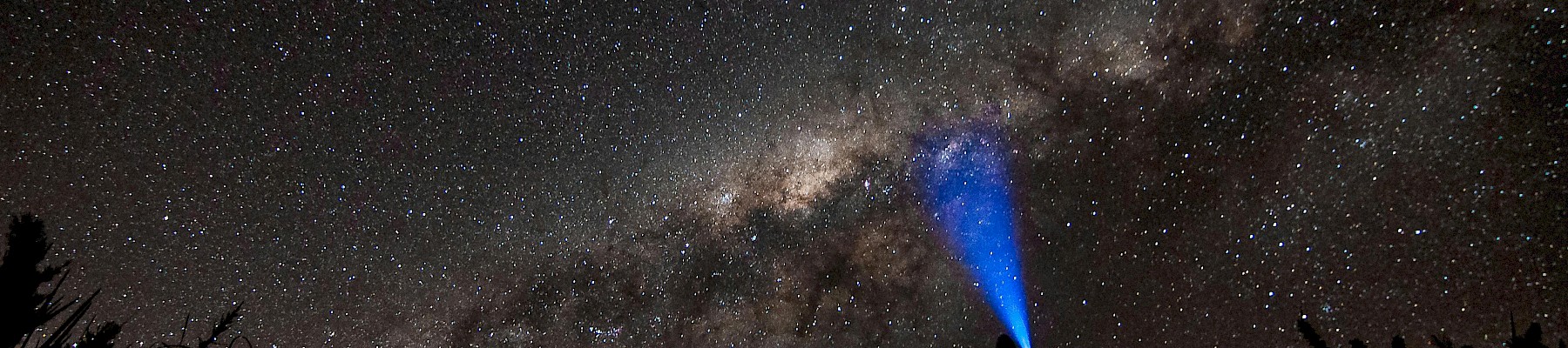 A person with a flashlight under a starry night sky, pointing towards the Milky Way galaxy in a silhouette view.