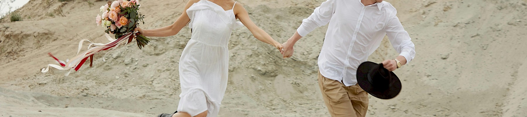 A couple is running hand-in-hand across a sandy desert landscape, the woman holding a bouquet and both smiling happily.