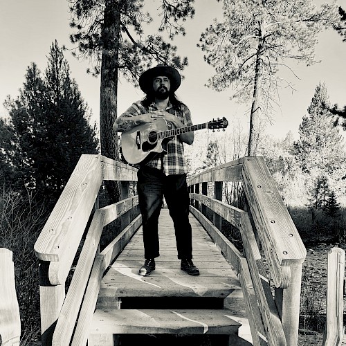 Dom Ruben stands on a wooden bridge holding an acoustic guitar among Central Oregon pine trees, promoting live music performances at Currents Restaurant & Lounge at Riverhouse Lodge in Bend, Oregon.