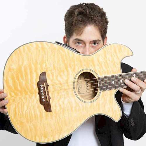 A man holds an acoustic guitar partially hiding his face, looking directly at the camera, against a plain, light background.