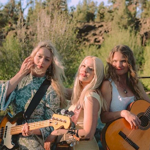 Three women with guitars pose by a river with rocks and greenery, enjoying a sunny day outdoors.