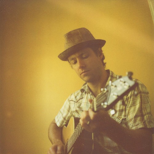 A man wearing a hat is playing an acoustic guitar against a yellow wall.