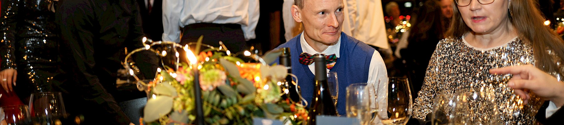 People sit and stand around a decorated table at a festive dinner event, with a mix of attentive, talking, and standing individuals.