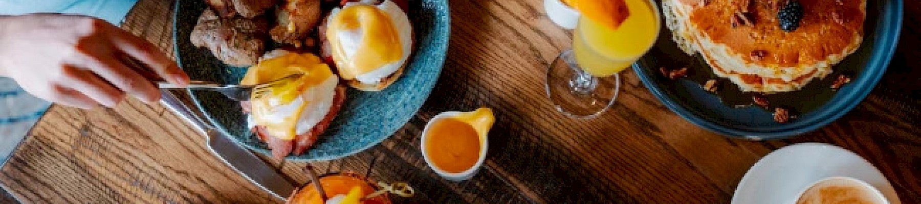 People are enjoying brunch with eggs Benedict, coffee, juice, and a stack of pancakes at a rustic wooden table.