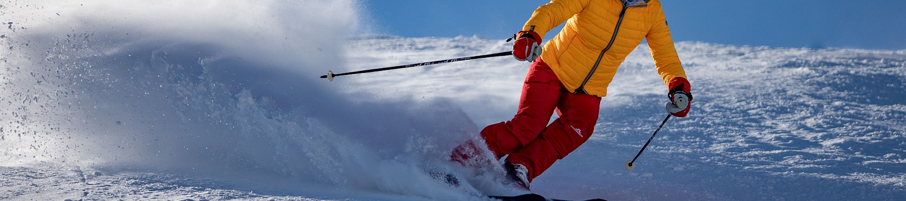 A skier in a yellow jacket and red pants winds down a snowy mountain slope under a clear blue sky.