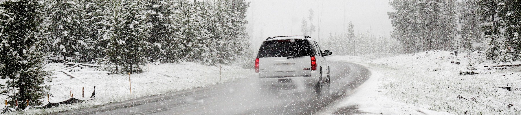 A car drives on a snowy, winding road surrounded by snow-covered trees during a winter storm.