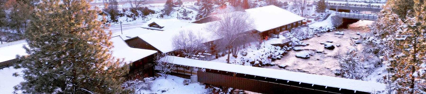 A snowy landscape features a river, a covered bridge, buildings, and trees, with the sun setting in the background.
