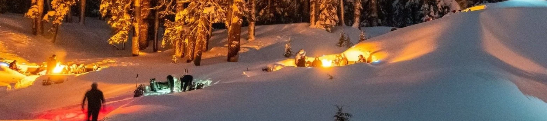 People clearing snow at night with orange portable lights glowing amid snowbanks and trees, a small group gathered around a snow pit.