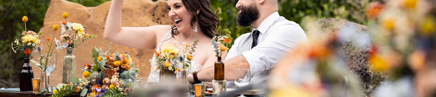 A smiling couple toasting at a decorated outdoor wedding reception, colorful floral arrangements, warm lanterns, and a rustic long table.