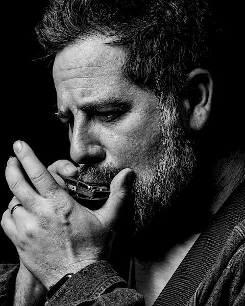 Black-and-white portrait of a singer-songwriter playing harmonica and acoustic guitar during a live performance at Currents at Riverhouse Lodge, a riverfront live music and dining venue in Bend, Oregon.