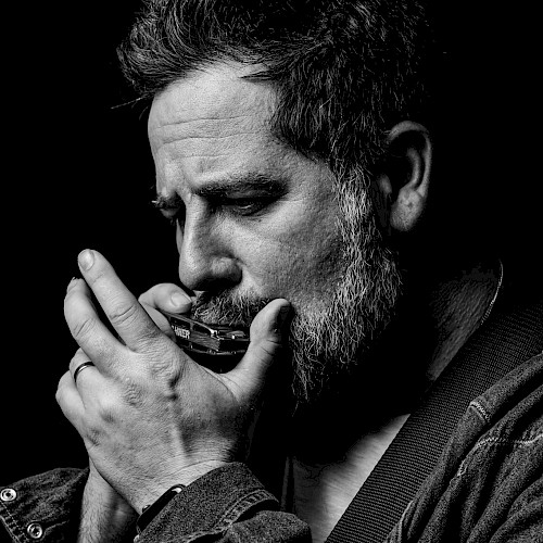 Black-and-white portrait of a singer-songwriter playing harmonica and acoustic guitar during a live performance at Currents at Riverhouse Lodge, a riverfront live music and dining venue in Bend, Oregon.