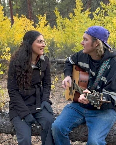 Two musicians sit outdoors on a fallen log surrounded by yellow foliage. A man wearing a backward cap plays an acoustic guitar while singing, facing a woman in a dark jacket who sings along, creating an intimate live-music moment in a forest setting.
