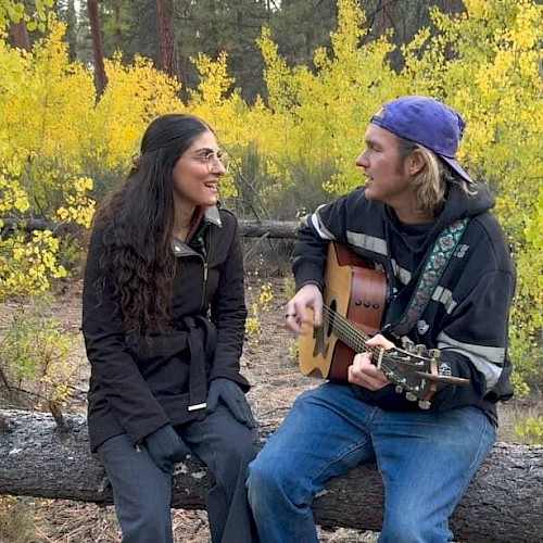 Two musicians sit outdoors on a fallen log surrounded by yellow foliage. A man wearing a backward cap plays an acoustic guitar while singing, facing a woman in a dark jacket who sings along, creating an intimate live-music moment in a forest setting.