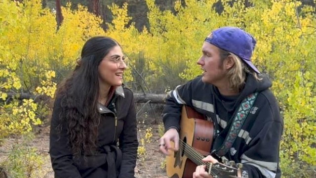 Two musicians sit outdoors on a fallen log surrounded by yellow foliage. A man wearing a backward cap plays an acoustic guitar while singing, facing a woman in a dark jacket who sings along, creating an intimate live-music moment in a forest setting.