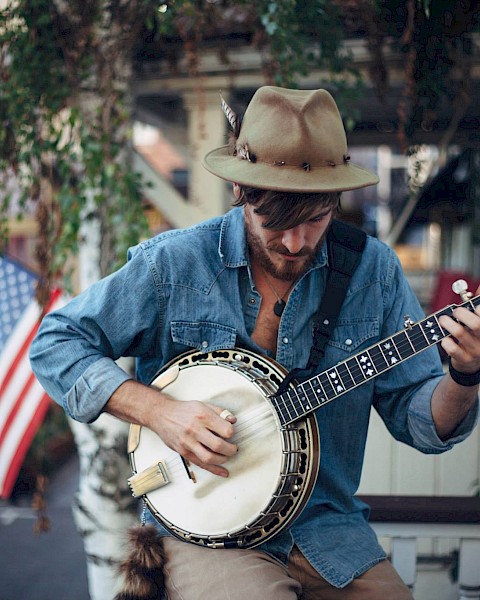 A solo musician wearing a wide-brim hat plays a banjo while seated outdoors, focused on the instrument. An American flag hangs in the background near a porch, with greenery and a neighborhood street visible behind him.
