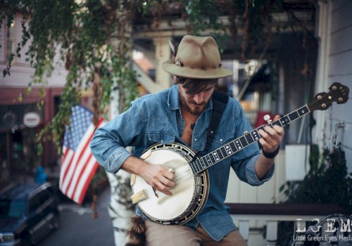 A solo musician wearing a wide-brim hat plays a banjo while seated outdoors, focused on the instrument. An American flag hangs in the background near a porch, with greenery and a neighborhood street visible behind him.