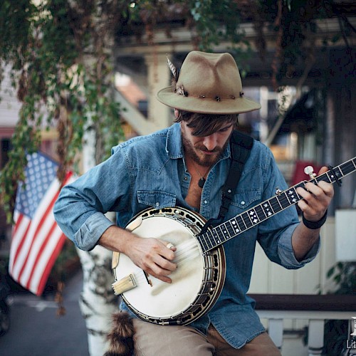 A solo musician wearing a wide-brim hat plays a banjo while seated outdoors, focused on the instrument. An American flag hangs in the background near a porch, with greenery and a neighborhood street visible behind him.