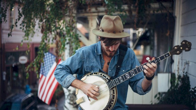 A solo musician wearing a wide-brim hat plays a banjo while seated outdoors, focused on the instrument. An American flag hangs in the background near a porch, with greenery and a neighborhood street visible behind him.