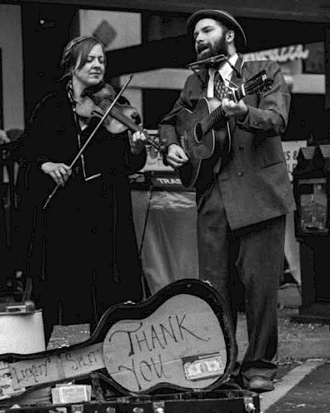 Black-and-white photo of a roots music duo performing live outdoors. A woman plays fiddle while singing, and a man sings while playing acoustic guitar and harmonica. An open guitar case with a “Thank You” sign sits in front, evoking old-time folk, jug band, and Americana street-style performance.