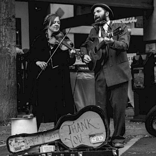 Black-and-white photo of a roots music duo performing live outdoors. A woman plays fiddle while singing, and a man sings while playing acoustic guitar and harmonica. An open guitar case with a “Thank You” sign sits in front, evoking old-time folk, jug band, and Americana street-style performance.