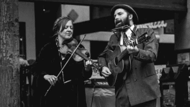 Black-and-white photo of a roots music duo performing live outdoors. A woman plays fiddle while singing, and a man sings while playing acoustic guitar and harmonica. An open guitar case with a “Thank You” sign sits in front, evoking old-time folk, jug band, and Americana street-style performance.