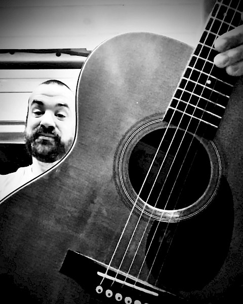A person takes a selfie with an acoustic guitar, showing part of the guitar's body and fretboard in a black-and-white photo.