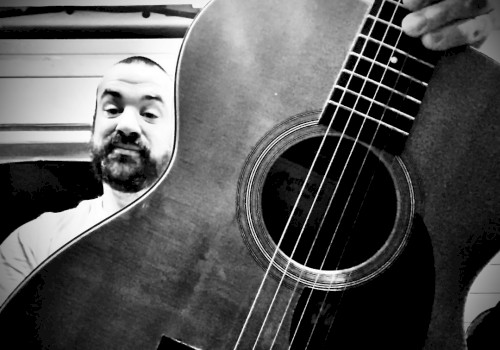 A person takes a selfie with an acoustic guitar, showing part of the guitar's body and fretboard in a black-and-white photo.