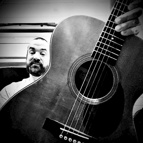 A person takes a selfie with an acoustic guitar, showing part of the guitar's body and fretboard in a black-and-white photo.