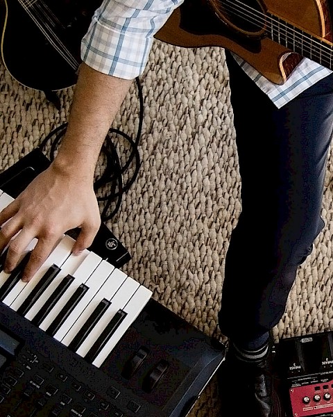 A musician is playing electronic and acoustic instruments, including a keyboard, guitar, and drum machine, on a carpeted floor.