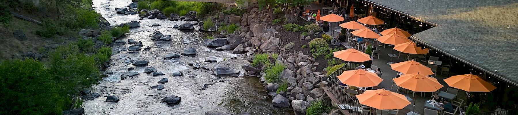 A riverside restaurant with outdoor seating and orange umbrellas next to a flowing, rocky creek surrounded by trees.