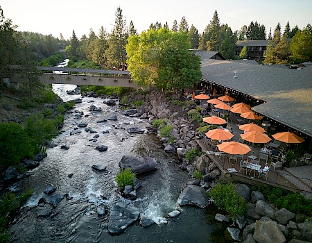 A riverside restaurant with outdoor seating and orange umbrellas next to a flowing, rocky creek surrounded by trees.