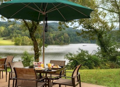 Outdoor patio with a table set for a meal, under a green umbrella, overlooking a serene lake and lush greenery in the background.