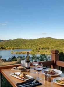 A dining table on a balcony features plates of food, wine, utensils, and glasses, overlooking a scenic view of a lake and lush, green hills.