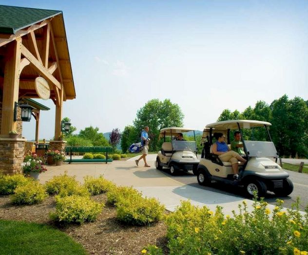 Two golf carts with drivers are parked near a building; one person is walking away carrying golf clubs. Sunny day with greenery around.