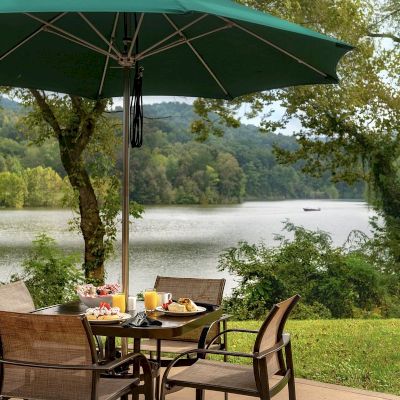 A lakeside outdoor dining area with a table set for breakfast under a green umbrella, surrounded by nature with a scenic lake view.