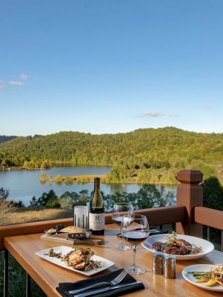 A wooden table set with meals and wine overlooks a scenic view of a lake surrounded by lush green hills, under a clear blue sky.