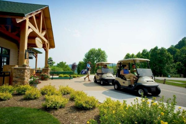 People with golf carts outside a building surrounded by greenery, with a person carrying golf bags. The scene is bright and clear, during the day.