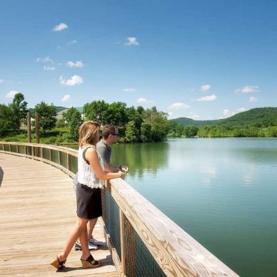 Two people stand on a wooden boardwalk overlooking a calm lake, surrounded by greenery and hills under a clear blue sky with a few clouds.