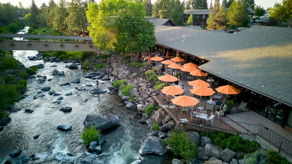 A riverside restaurant with orange umbrellas and outdoor seating, surrounded by trees and rocks, overlooking a flowing creek.