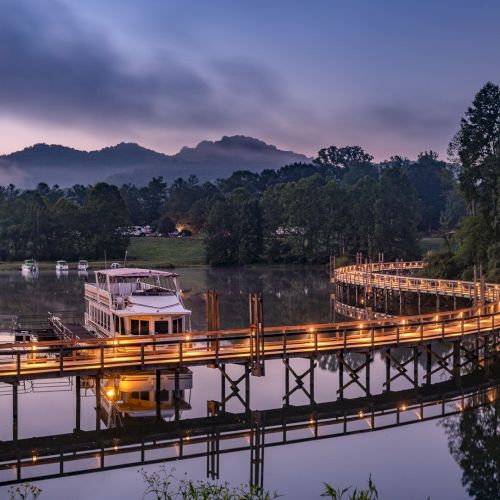 A serene lakeside scene with a boat docked at a glowing, curved wooden bridge set against a backdrop of mountains and trees at twilight.