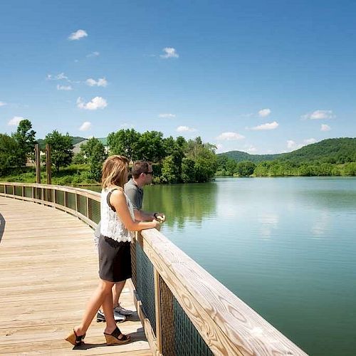 Two people are standing on a wooden boardwalk overlooking a peaceful lake surrounded by lush green hills and clear blue skies.