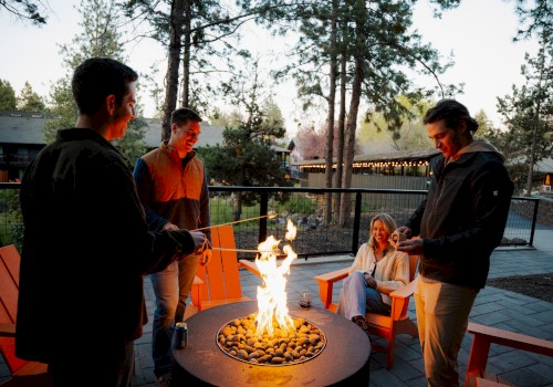 Four friends enjoy a campfire outdoors at dusk, sitting and standing around, with smiles and laughter.